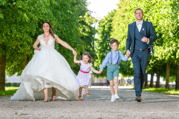 mariés et leurs enfants courant dans une allée bordée d'arbres