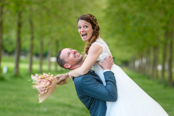 couple de mariés le marié porte sa femme devant une rangée d'arbres