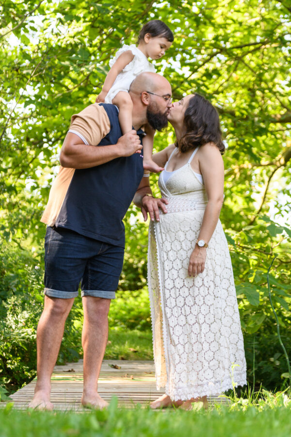 portrait femme enceinte embrassant son mari dans la nature, un enfant posé sur les épaules du père les regardant
